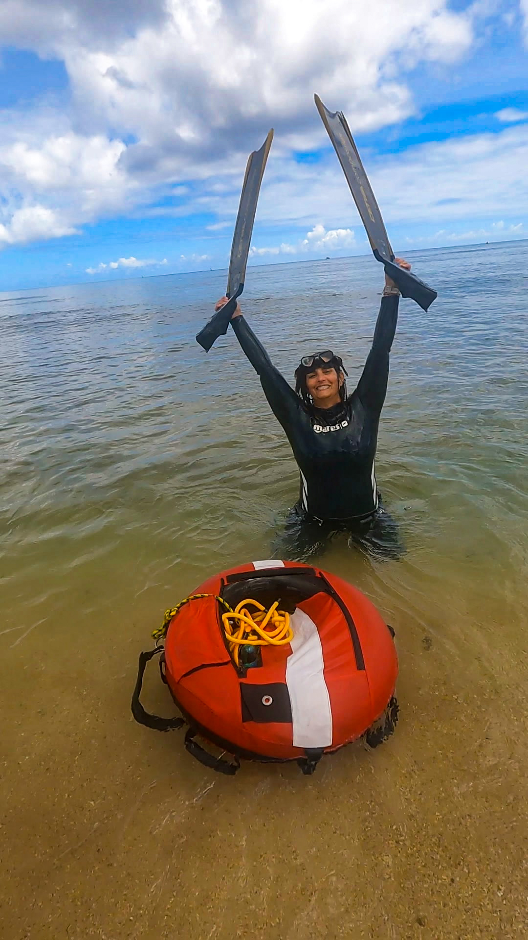 Elaine Hicks smiling in shallow water with her freediving fins held overhead.